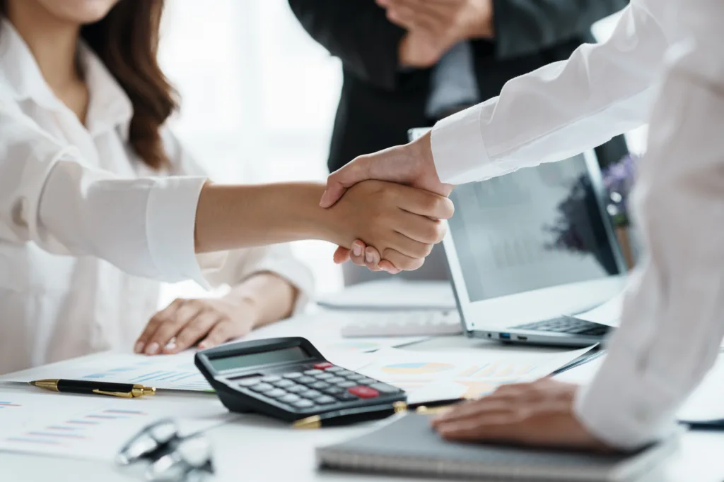 Business professionals shaking hands during a financial meeting with reports and a calculator on the desk.