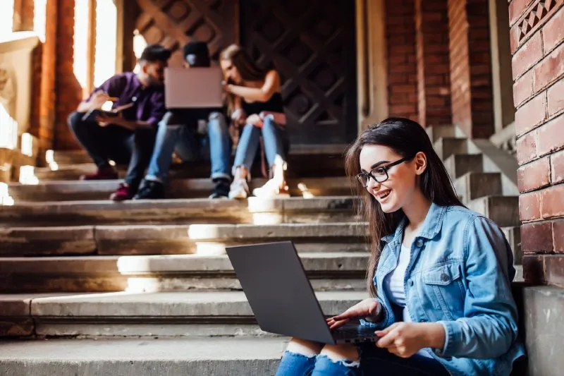 College student using a laptop to access their student loan portal on a stairway on campus