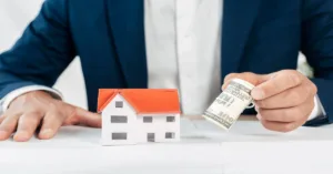 Hand holding cash near house model representing hard money loan requirements