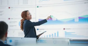 A professional woman in a suit pointing to a line graph on a large digital display while holding a tablet, presenting data to a team.