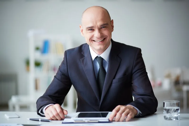Smiling businessman in dark suit sitting at desk with tablet, pen, and water glass.