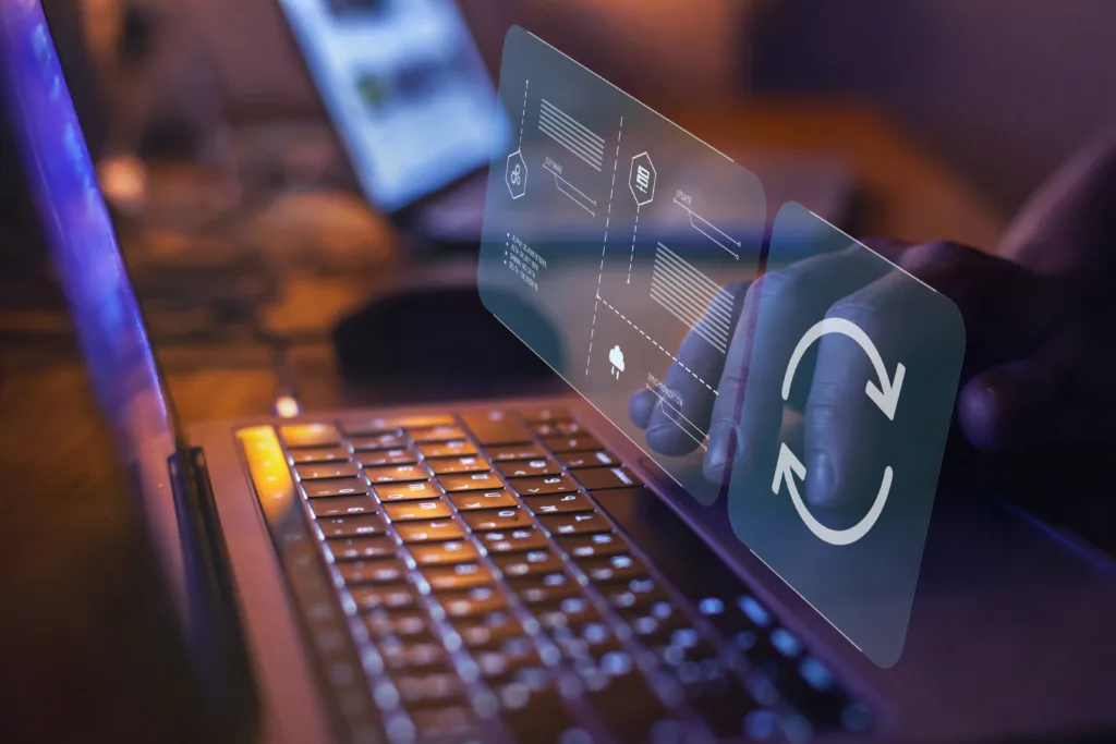 A hand on a laptop trackpad with glowing, holographic data icons and a sync symbol floating above the keyboard.