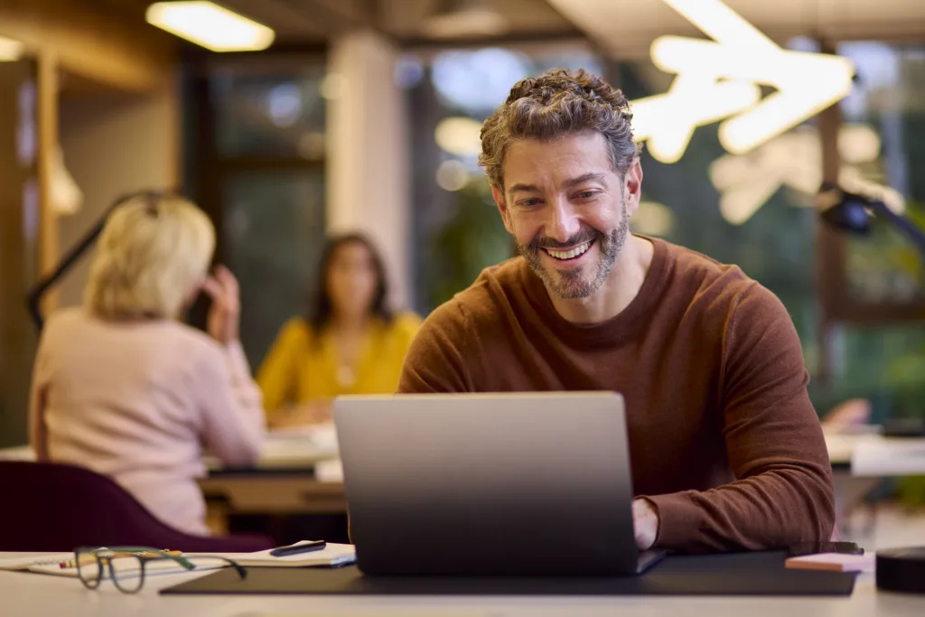 A man smiling while working on a laptop in a bright, modern office setting.
