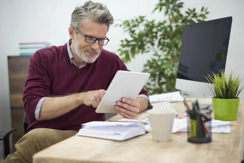 Man in maroon sweater using a tablet at a desk with a computer and plant.