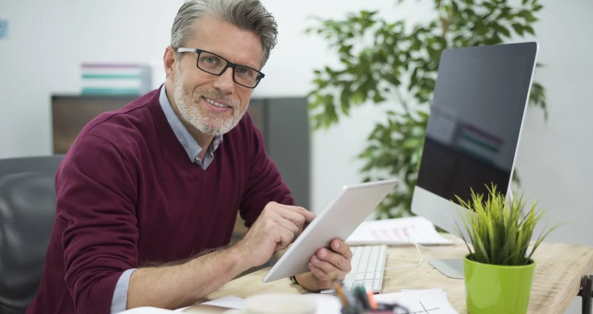 Smiling man in glasses holding a tablet at a desk with a computer and plant.