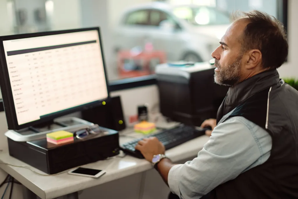 Auto shop manager reviewing loan or inventory data on a desktop computer.