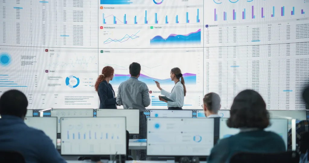 Three professionals standing in a high-tech command center, discussing complex data analytics displayed on a massive, multi-panel wall screen.