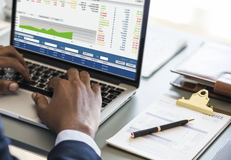 Close-up of hands typing on laptop displaying stock trading charts, invoice and pen nearby.