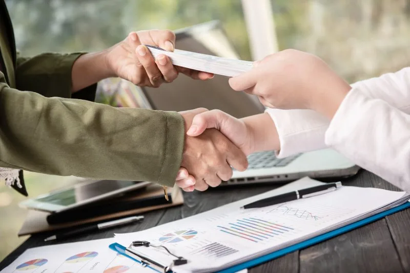 Two banking professionals shaking hands while exchanging loan documents over portfolio reports. 
